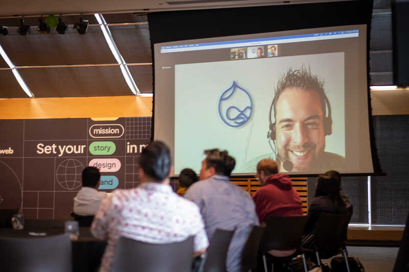 Dries Butaert appears on a large screen for a Q&A session at the EvolveDrupal summit. He is smiling, looking at a room of people sitting around tables.