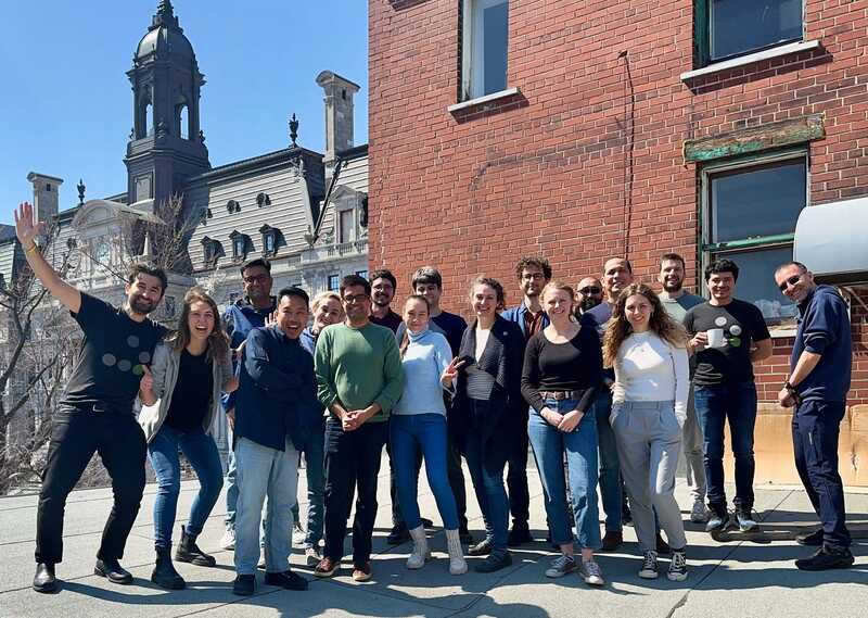 Eighteen Evolving Web team members stand together and smile at the camera on a flat rooftop in Montreal on a sunny day. 