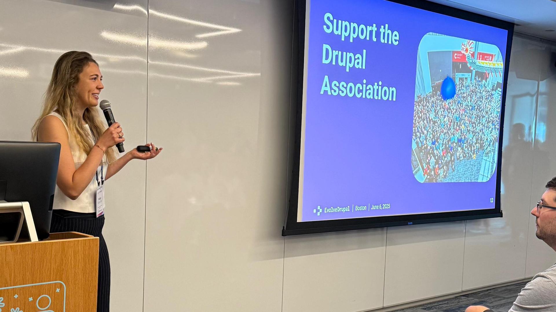 Maya with a microphone standing in front of a screen saying Support the Drupal Association