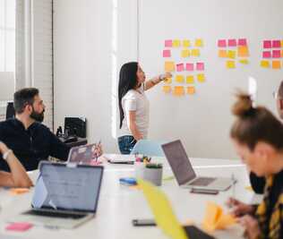 A group of young people sitting around a table in a meeting room with their laptops looking at a woman pointing at a post it on a wall