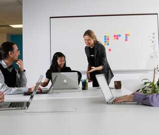 Several people are sitting staring at a computer screen while Suzanne is standing and presenting