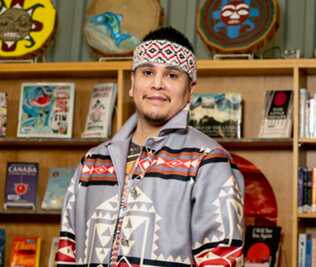A young man standing before a shelf in traditional Indigenous attire