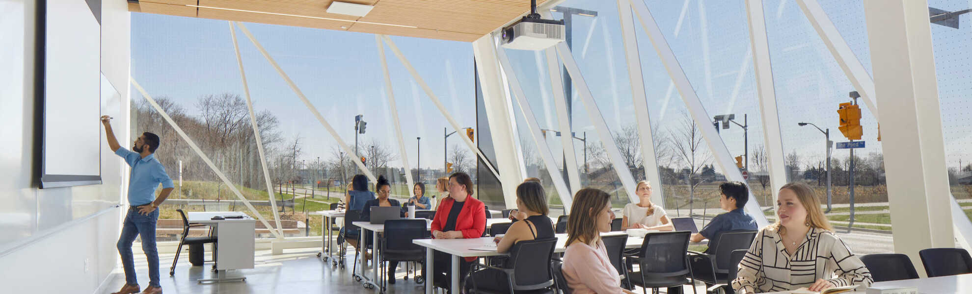 A photo banner of a bright classroom with glass walls with a person at the whiteboard on the left and students sitting at a row of desks on the right.