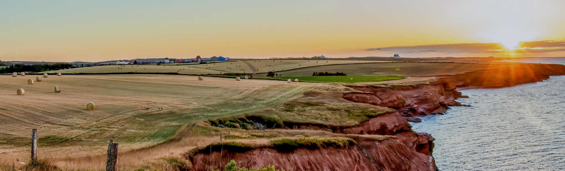 Scenic view of Prince Edward Island’s red sandstone cliffs at sunset, with farmland and hay bales stretching into the distance beside the ocean