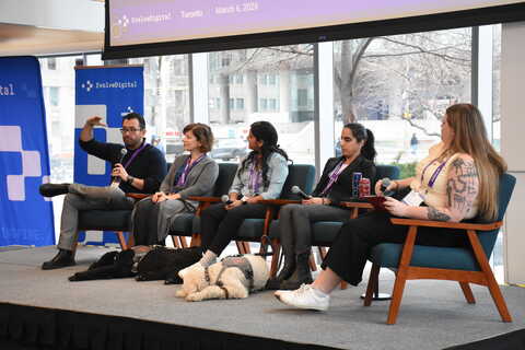 Five panelists speaking on an accessibility panel at EvolveDigital Toronto 2026, seated on stage with microphones, while two service dogs lie calmly at their feet.