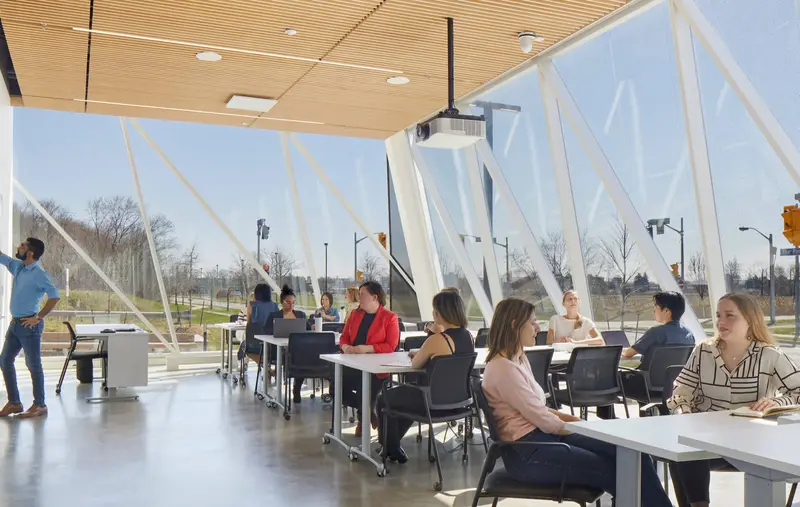 A photo banner of a bright classroom with glass walls with a person at the whiteboard on the left and students sitting at a row of desks on the right.