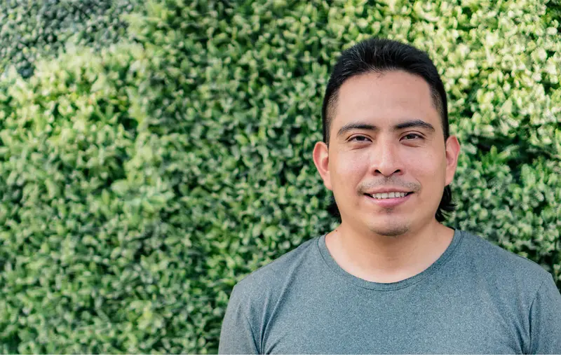 A portrait of a person facing the camera and smiling against some bright green bushes.