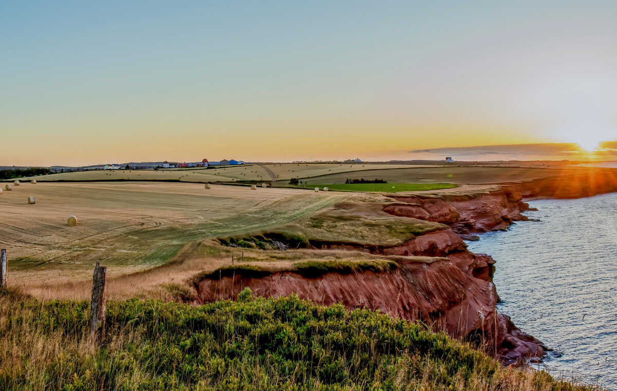 Scenic view of Prince Edward Island’s red sandstone cliffs at sunset, with farmland and hay bales stretching into the distance beside the ocean