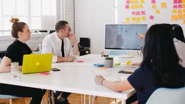 3 people in a meeting room looking a computer screen on a table.
