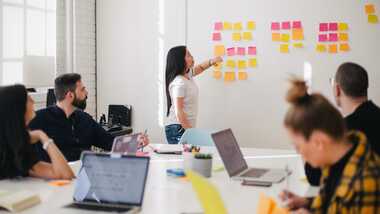 A group of young people sitting around a table in a meeting room with their laptops looking at a woman pointing at a post it on a wall